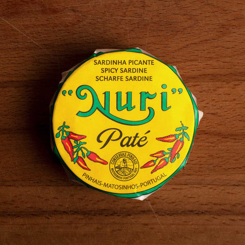 A round tin in a yellow paper wrap. Nuri is written in green lettering above some drawings of red chilli peppers.