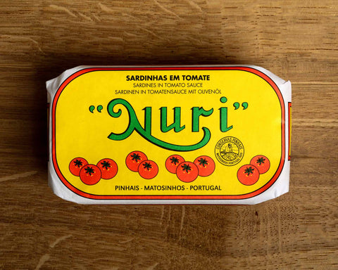 A tin of sardines in a yellow paper wrap with the Nuri logo in green on the front above some images of tomatoes. The tin has a red and white border and is on a wood backdrop.