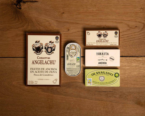 A selection of four small tins and one large of Cantabrian anchovies in classic packaging. The tins are against a wooden backdrop.