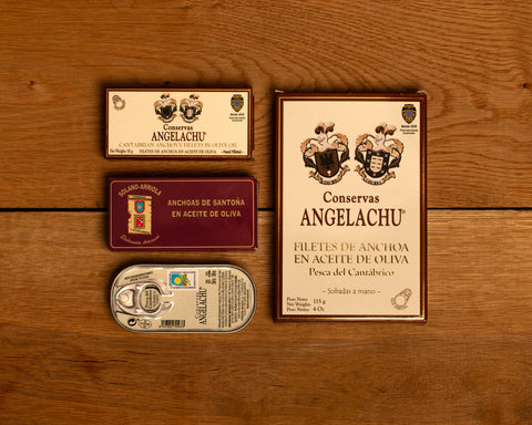 A selection of three smaller tins next to one larger tin of Cantabrian anchovies, all in classic packaging, against a wood background.