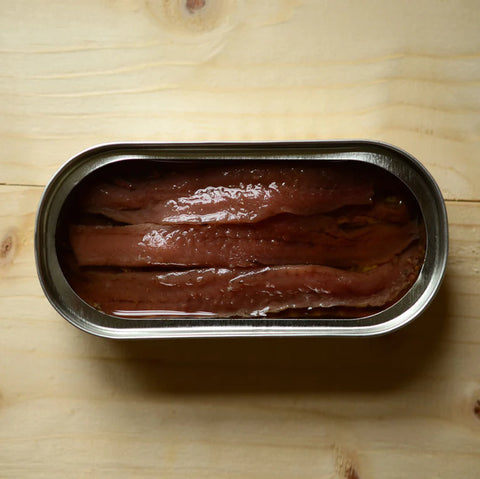 An open tin of anchovies in a silver trimmed tin resting on a light brown table.