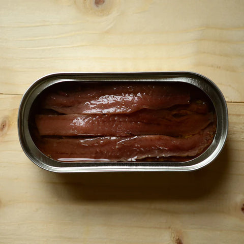 An open tin of anchovies in a silver trimmed tin resting on a light brown table.