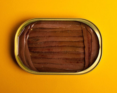 An open tin of anchovy fillets. The long, rich brown fillets are framed by shorter fillets at either end of the tin. The tin is on a bright yellow background