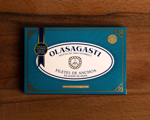 A tin of large anchovy fillets in blue and white packaging against a wood background