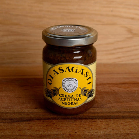A glass jar of tapenade with a yellow label on the front depicting the Olasagasti logo and images of black olives