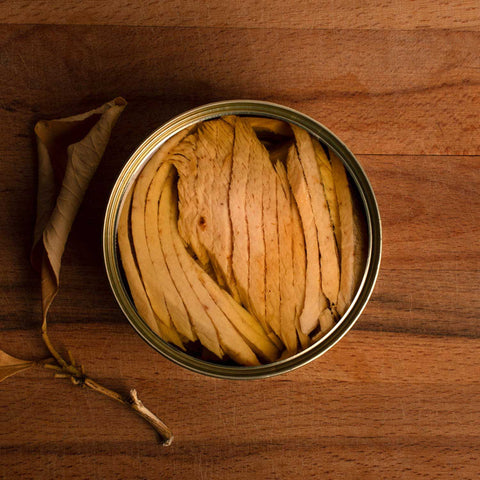 A round tin of finely sliced tuna belly in olive oil by a dried leaf on a wood background