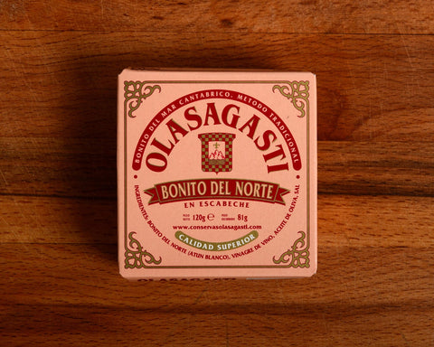 A tin of Bonito del Norte tuna in pink packaging with red lettering and an ornate gold border, against a wood background