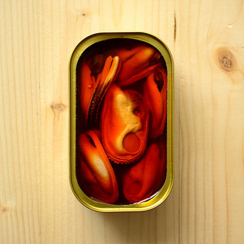 An open tin of large mussels in orange escabeche. The tin is on a pale pine wood background