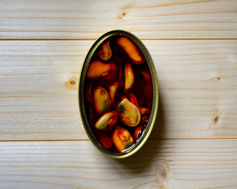 An open, oval-shaped tin of orange mussels in escabeche against a pale pine wood background