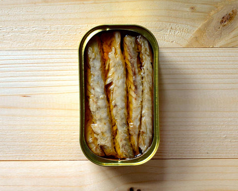 An open tin of mackerel fillets in spicy olive oil against a pale pine wood background.