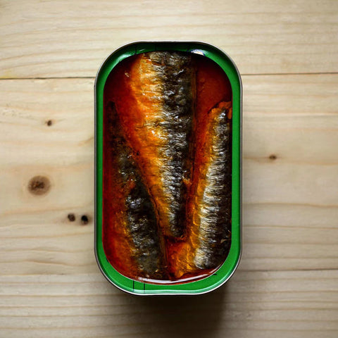 Three sardines in a rich red sauce in an open tin with a green trim against a pale pine table.