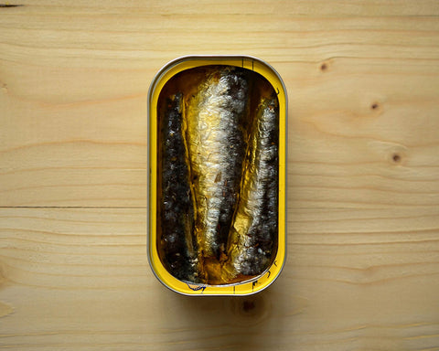 An open tin of sardines in a golden-coloured olive oil against a pale pine background.