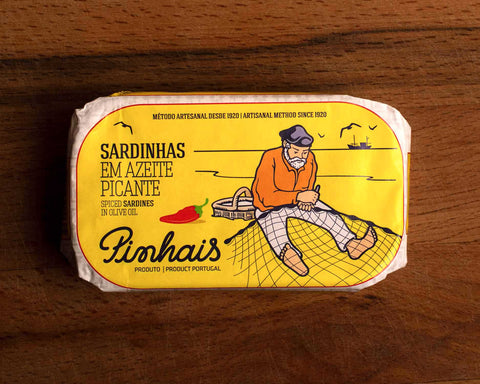 A tin of spicy sardines in a yellow paper wrap with an image of a fisherman mending a fishing net on the front next to a red chilli pepper. The tin is on a wood background.