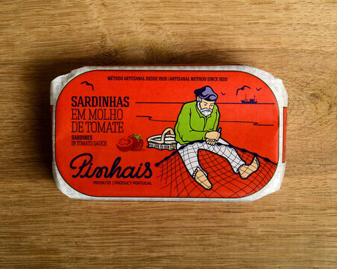 A tin of sardines in a red paper wrap with a white border. A fisherman in a green jersey mends his net on the front. The tin is on a wood background.