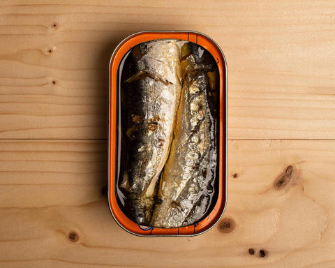 An open tin of skin-on sardines in olive oil against a pale pine wood background.