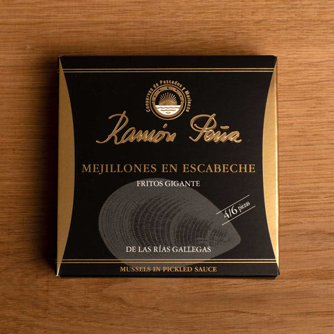 A tin in square black card packaging. Ramón Peña is written in gold lettering above an image of a silvery grey mussel. The tin is on a wood background.