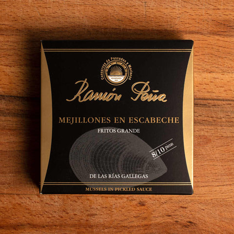 A tin of mussels in black card packagaing with a gold border and an image of a mussel drawn on grey on the front. The tin is on a wood background. 