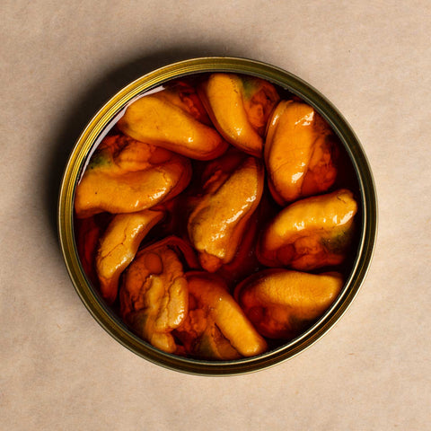 An open tin of large Galician mussels. The tin is round and the mussels are bright orange and arranged in a circle inside the tin. The tin is against a pale background.
