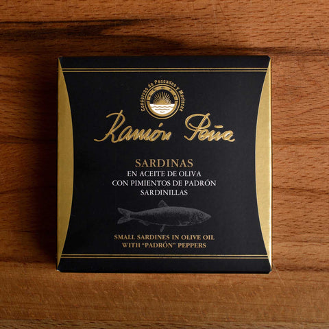 A tin of sardines in a square black and gold card box. Ramón Peña is written in gold lettering above a silver-grey image of a sardine.