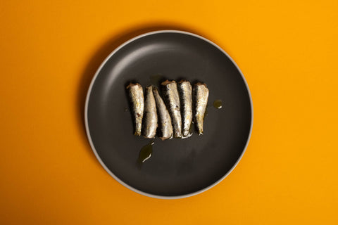 Six small sardines arranged in a line on a round grey plate against a yellow background.