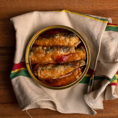 An open tin of sardines, lightly battered and in a sauce. The tin is resting on a crumpled tea towel with red, green and yellow stripes.