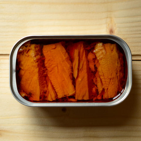 An open tin of four tuna fillets in a bright orange oil, against a pale pine background.