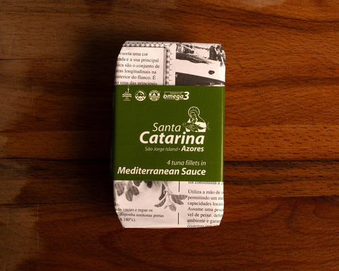 A tin of Santa Catarina tuna in newspaper-style packaging with a green label against a wood background