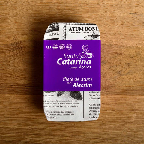A tin of Santa Catarina tuna in newspaper-style packaging with a purple label against a wood background