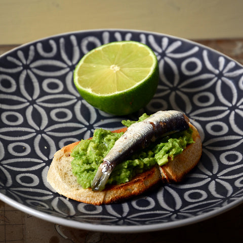 Mashed avocado on a slice of toasted bread topped with a sardine, on a patterned plate. 