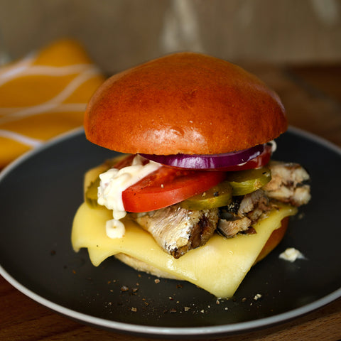 A close up of a sardine burger with a juicy patty topped with sliced tomato, jalapeno peppers, red onion and mayo, between two brioche buns, on a black plate with a thin white border.
