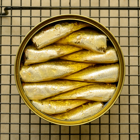 An open tin of sardine fillets in extra virgin olive oil against a steel cage.