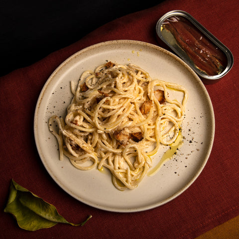 A plate of spaghetti with an open tin of anchovy fillets. Delicious combination of flavours and textures. Presented on a cream circular plate resting on a wine table cloth.