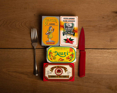 Four brightly coloured tins of fish with a fork and a red knife on either side, against a wood background.