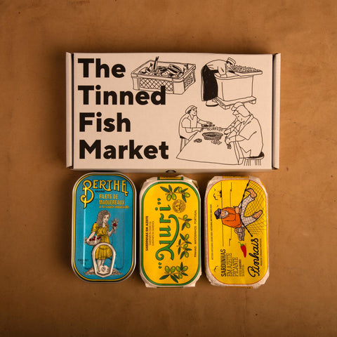 Three colourfully packaged tins of fish beneath a gift box illustrated with line drawings of the cannery workers, a basket of fish and The Tinned Fish Market in bold black lettering.
