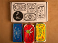 Three colourfully packaged tins of fish beneath a gift box illustrated with line drawings of the tins and The Tinned Fish Market logo.