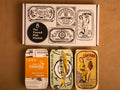Three colourfully packaged tins of fish beneath a gift box illustrated with line drawings of the tins and The Tinned Fish Market logo.