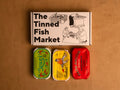 Three colourfully packaged tins of fish beneath a gift box illustrated with line drawings of the cannery workers, a basket of fish and The Tinned Fish Market in bold black lettering.