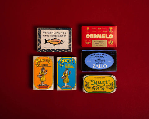 A selection of colourfully-packaged tinned 
sardines, tuna, mackerel, salmon and carmelo sardines against a red background.