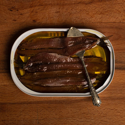 An open tin of butterfly anchovies in olive oil. A small silver spoon is fishing one of the anchovies from the tin. The tin is on a light brown wooden counter. 