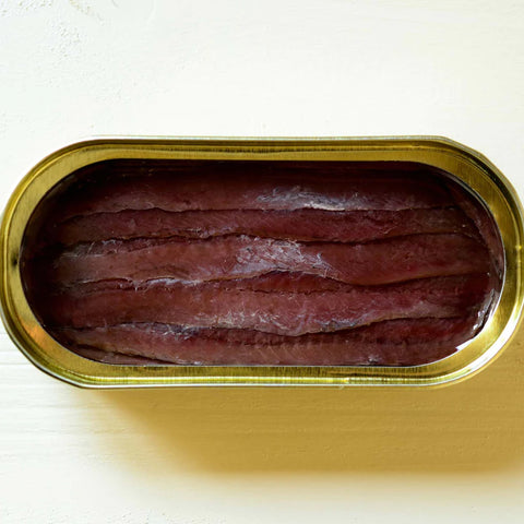 Tinned reddish-brown anchovies in a tin with gold trim placed upon a light wooden table.